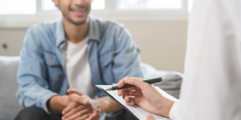 Doctor in white coat taking notes while consulting with smiling male patient during medical evaluation for car accident or workers’ compensation injury
